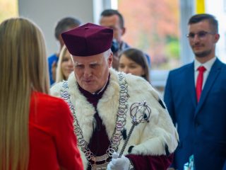 Rektor w uroczystym stroju stoi przed studentką w czerwonej bluzce podczas immatrykulacji.