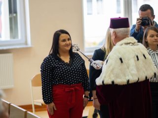 Rektor dotyka berłem ramienia studentki w bluzce w kropki i czerwonych spodniach.