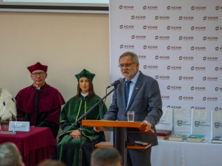 Mężczyzna w okularach i szarym garniturze przemawia przy mównicy.