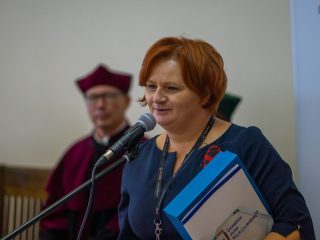 Kobieta z krótkimi rudymi włosami przemawia, trzymając teczki z dokumentami.