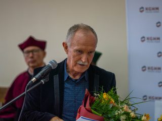 Mężczyzna z siwym wąsem i bukietem kwiatów stoi przy mikrofonie.