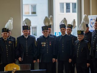 Chórzyści w mundurach górniczych i czakach z białymi pióropuszami stoją w szeregu.