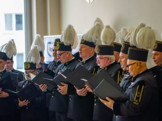 Chór górniczy śpiewa, trzymając otwarte czarne teczki z nutami.