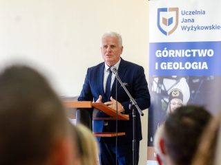 Siwowłosy mężczyzna przemawia przy mównicy obok banera kierunku Górnictwo i geologia.