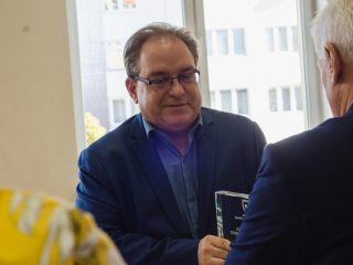 Mężczyzna w okularach odbiera szklaną nagrodę od przedstawiciela uczelni.