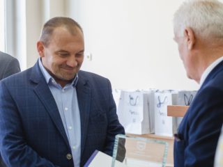 Uśmiechnięty mężczyzna w granatowej marynarce odbiera wyróżnienie od siwowłosego organizatora.