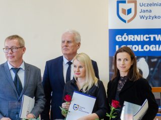 Mężczyzna i dwie kobiety prezentują dyplomy oraz nagrody obok siwowłosego przedstawiciela uczelni.