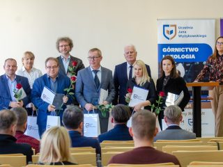 Nagrodzeni pozują przed publicznością, a prowadząca stoi przy mównicy.