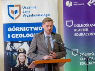 Mężczyzna w szarym garniturze przemawia przy mównicy na tle banerów uczelni i klastra edukacyjnego.