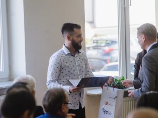Brodaty mężczyzna w jasnej wzorzystej koszuli odbiera dokument i upominek.