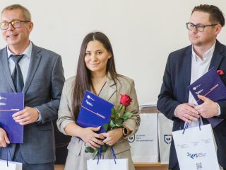 Kobieta w szarej sukience pozuje z czerwoną różą i teczką między dwoma mężczyznami.