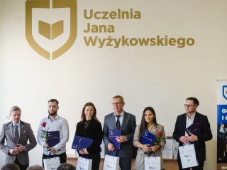 Nagrodzeni i organizator stoją pod dużym logo Uczelni Jana Wyżykowskiego.