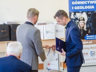 Mężczyzna w niebieskim garniturze odbiera torbę upominkową, trzymając fioletową teczkę.