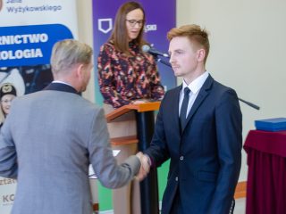 Młody mężczyzna w ciemnym garniturze ściska dłoń wręczającego nagrody.