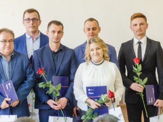 Kobieta i pięciu mężczyzn prezentują dokumenty, róże oraz torby upominkowe.