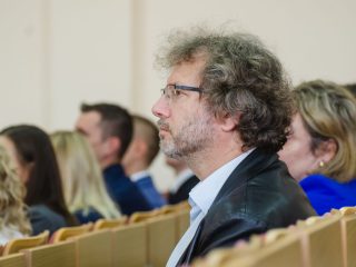 Mężczyzna w okularach i skórzanej kurtce słucha wystąpienia wśród publiczności.