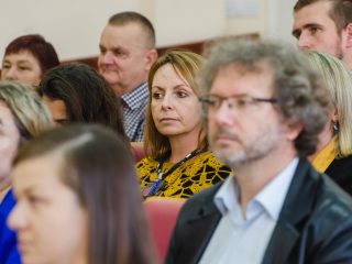 Goście uroczystości, w tym kobieta w żółtej wzorzystej bluzce, słuchają wystąpienia.