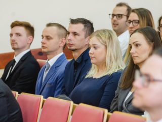 Kobiety i mężczyźni siedzą w rzędach czerwonych foteli podczas uroczystości.