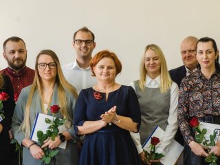 Organizatorka w granatowej sukience stoi wśród uśmiechniętych nagrodzonych z różami.