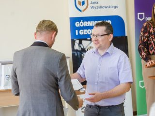 Mężczyzna w jasnofioletowej koszuli odbiera gratulacje od organizatora uroczystości.