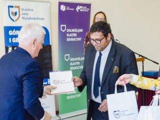 Mężczyzna w okularach i garniturze odbiera dyplom oraz białą torbę upominkową.