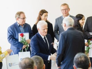 Przedstawiciel uczelni ściska dłoń uczestnika, a w tle stoją nagrodzeni z różami.