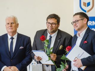 Siwowłosy przedstawiciel uczelni pozuje z dwoma nagrodzonymi trzymającymi dokumenty i róże.