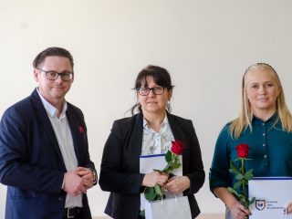 Mężczyzna w okularach pozuje z dwiema nagrodzonymi kobietami trzymającymi róże i dyplomy.