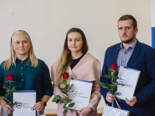 Troje nagrodzonych prezentuje dyplomy i czerwone róże na tle banera uczelni.