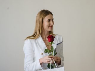 Kobieta w białej marynarce trzyma dyplom, czerwoną różę i torbę upominkową.