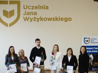 Grupa nagrodzonych pozuje z dokumentami i różami obok organizatorki.