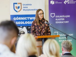 Prowadząca w okularach i wzorzystej bluzce przemawia przy mównicy.