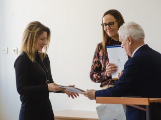 Kobieta w czarnej sukience odbiera dokument od siwowłosego przedstawiciela uczelni.