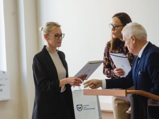 Kobieta w okularach i czarnej marynarce odbiera dyplom przy mównicy.