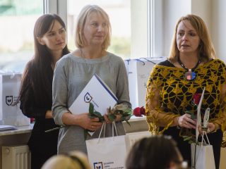 Nagrodzone kobiety stoją z różami, dyplomami i torbami przed oknami sali.