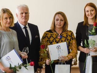 Trzy nagrodzone kobiety pozują z dyplomami i różami obok przedstawiciela uczelni.