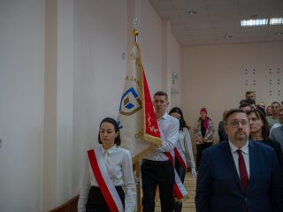 Poczet sztandarowy z biało-czerwonymi szarfami stoi ze sztandarem wśród uczestników uroczystości.