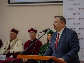 Mężczyzna w garniturze i czerwonym krawacie przemawia przy mównicy przed przedstawicielami uczelni.