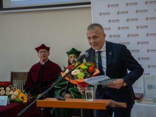 Mężczyzna z bukietem kwiatów i dokumentami przemawia przy mównicy.