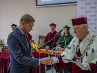 Mężczyzna w niebieskim garniturze odbiera szklaną statuetkę od rektora.