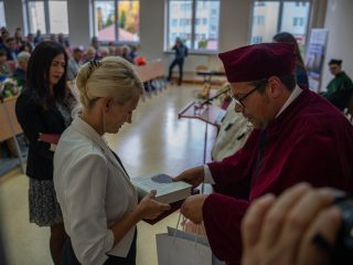Kobieta w białej bluzce odbiera książkę od przedstawiciela uczelni w bordowej todze.