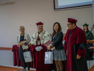 Dwie wyróżnione kobiety pozują z dokumentami i upominkami obok przedstawicieli uczelni w togach.