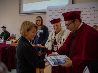 Kobieta odbiera książki od przedstawiciela uczelni w bordowej todze.