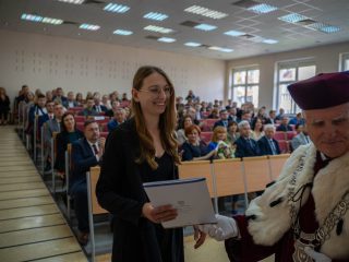 Uśmiechnięta kobieta w okularach trzyma dyplom obok rektora, z publicznością w tle.