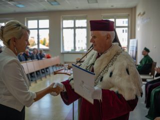 Kobieta w białej bluzce ściska dłoń rektora trzymającego teczkę z dokumentami.