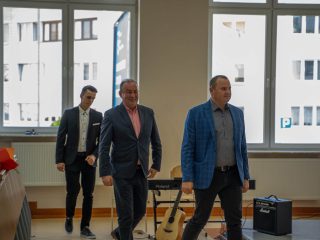 Trzej mężczyźni w garniturach stoją przed publicznością obok instrumentów muzycznych.