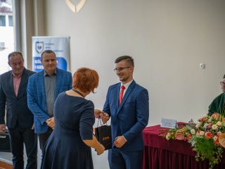 Kobieta z krótkimi rudymi włosami wręcza upominek mężczyźnie w niebieskim garniturze.