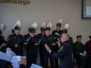 Dyrygent prowadzi chór w mundurach górniczych i czakach z białymi pióropuszami.