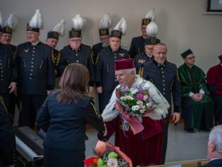 Rektor z bukietem kwiatów rozmawia z kobietą stojącą przed chórem górniczym.