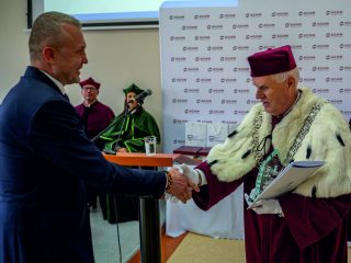 Mężczyzna w garniturze ściska dłoń rektora trzymającego dokumenty.